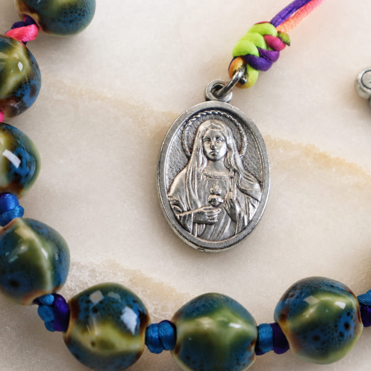 Sacred Heart medal attached to ceramic Catholic prayer beads with vibrant woven cord and handmade religious details.