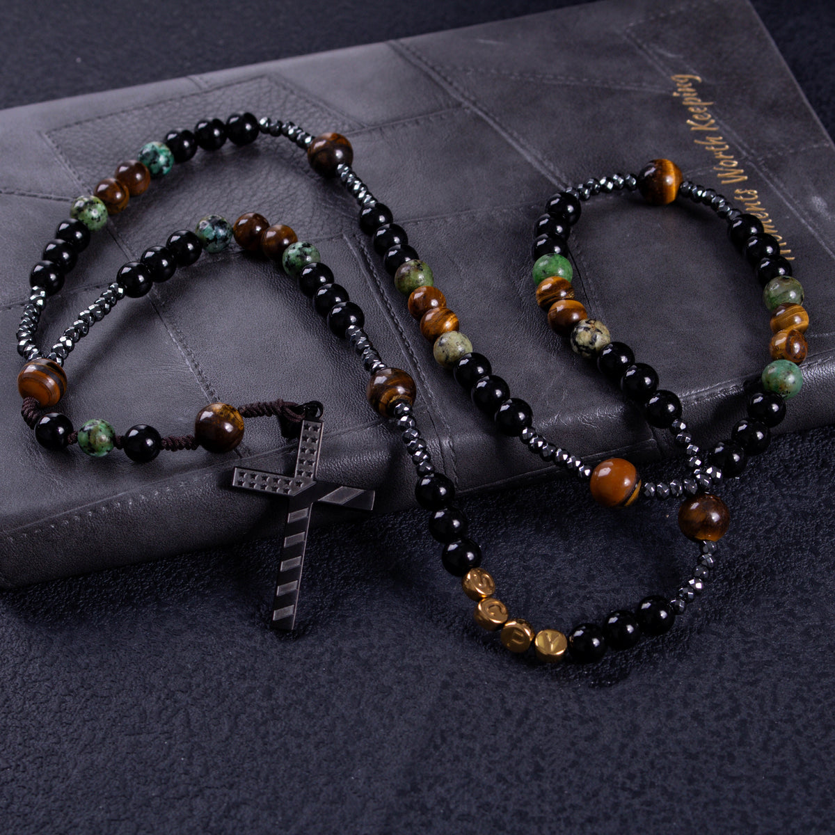 Stone Guardian Rosary featuring black prayer beads, earthy tiger-eye tones, green stone-style accents, and a bold black cross pendant.