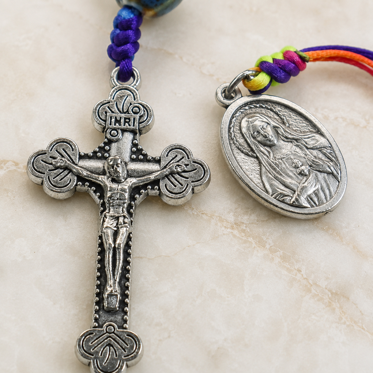 Close-up of ceramic-style Catholic prayer beads featuring blue green patterned beads, woven cord details, and religious medal.