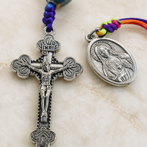 Close-up of ceramic-style Catholic prayer beads featuring blue green patterned beads, woven cord details, and religious medal.