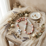 Custom name silicone rosary for baby on a warm wooden surface, surrounded by preserved flowers, letter beads spelling STEVIE MARI, boho earth-tone colors.