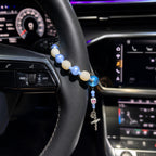 Blue Catholic car rosary displayed on a steering wheel with soft blue and white beads, personalized initial charm, Miraculous Medal, and silver crucifix