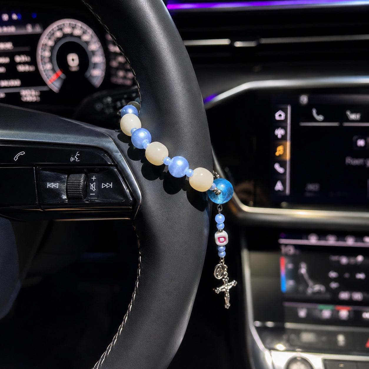 Blue Catholic car rosary displayed on a steering wheel with soft blue and white beads, personalized initial charm, Miraculous Medal, and silver crucifix