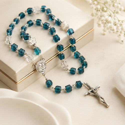 Blue Catholic rosary with crystal-style beads, Virgin Mary medal, and silver crucifix displayed on an elegant cream gift box