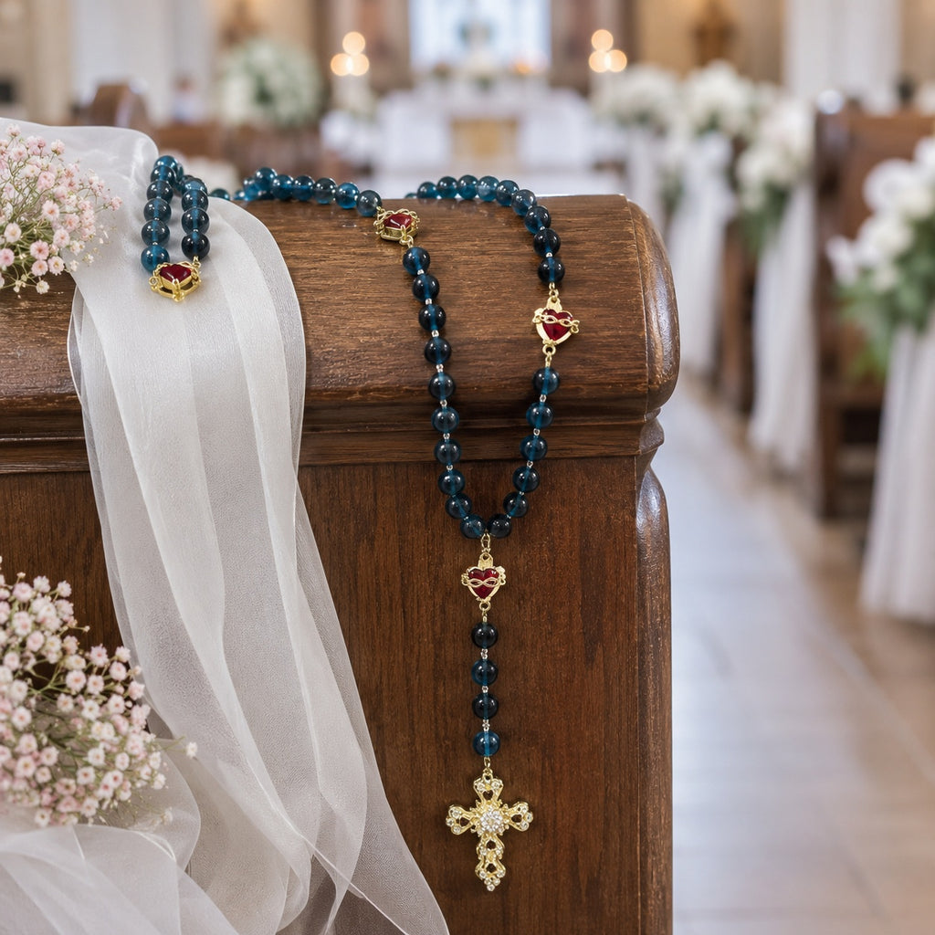 Elegant Catholic bridal rosary with blue-green beads, red heart accents, and ornate gold crucifix styled on wedding lace with rings and pearl earrings for a faith-filled ceremony detail.