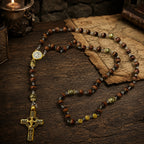 Vintage men’s prayer beads with brown gemstone-style beads, gold crucifix, and Catholic medal arranged on antique paper and dark wood.