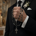 Men’s Catholic wedding rosary for groom with black and gray beads, silver crucifix, and Virgin Mary medal held with a formal suit