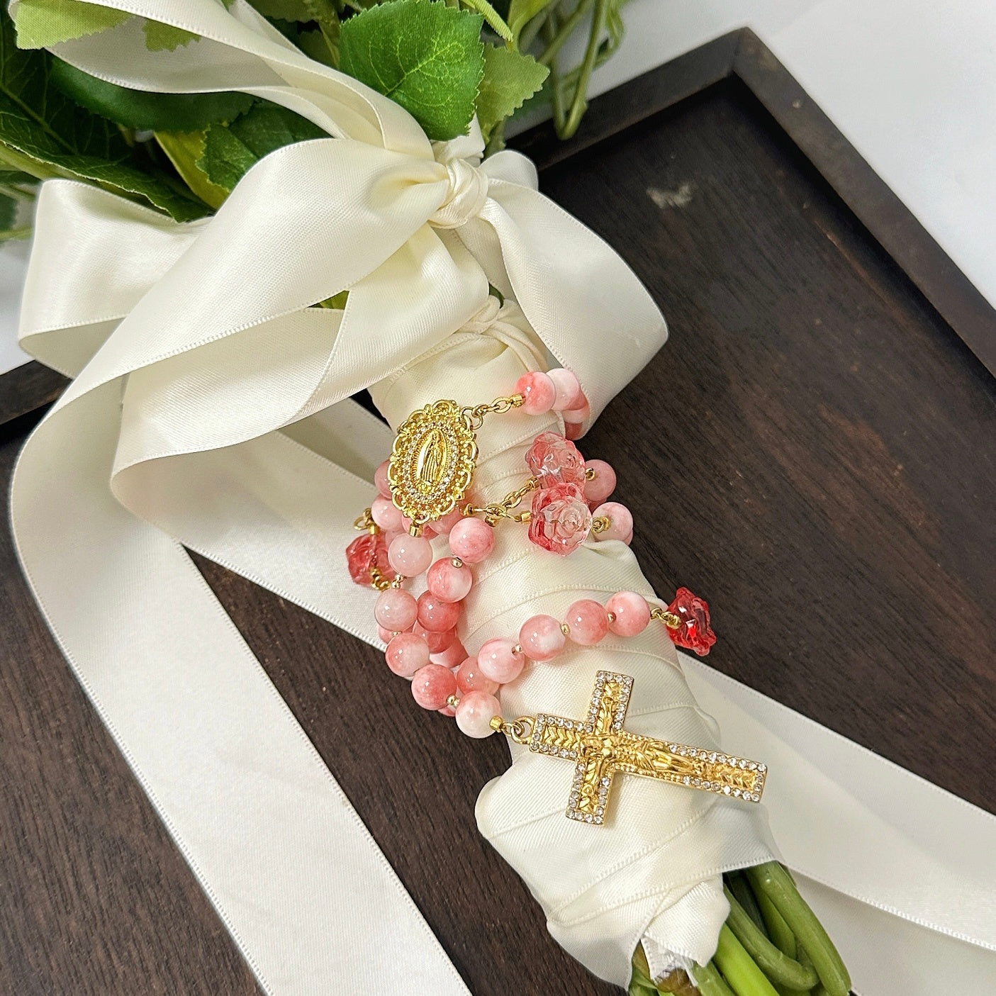 Pink Catholic wedding rosary for bride with gold crucifix, Marian medal, and rose accent beads styled on a bridal bouquet