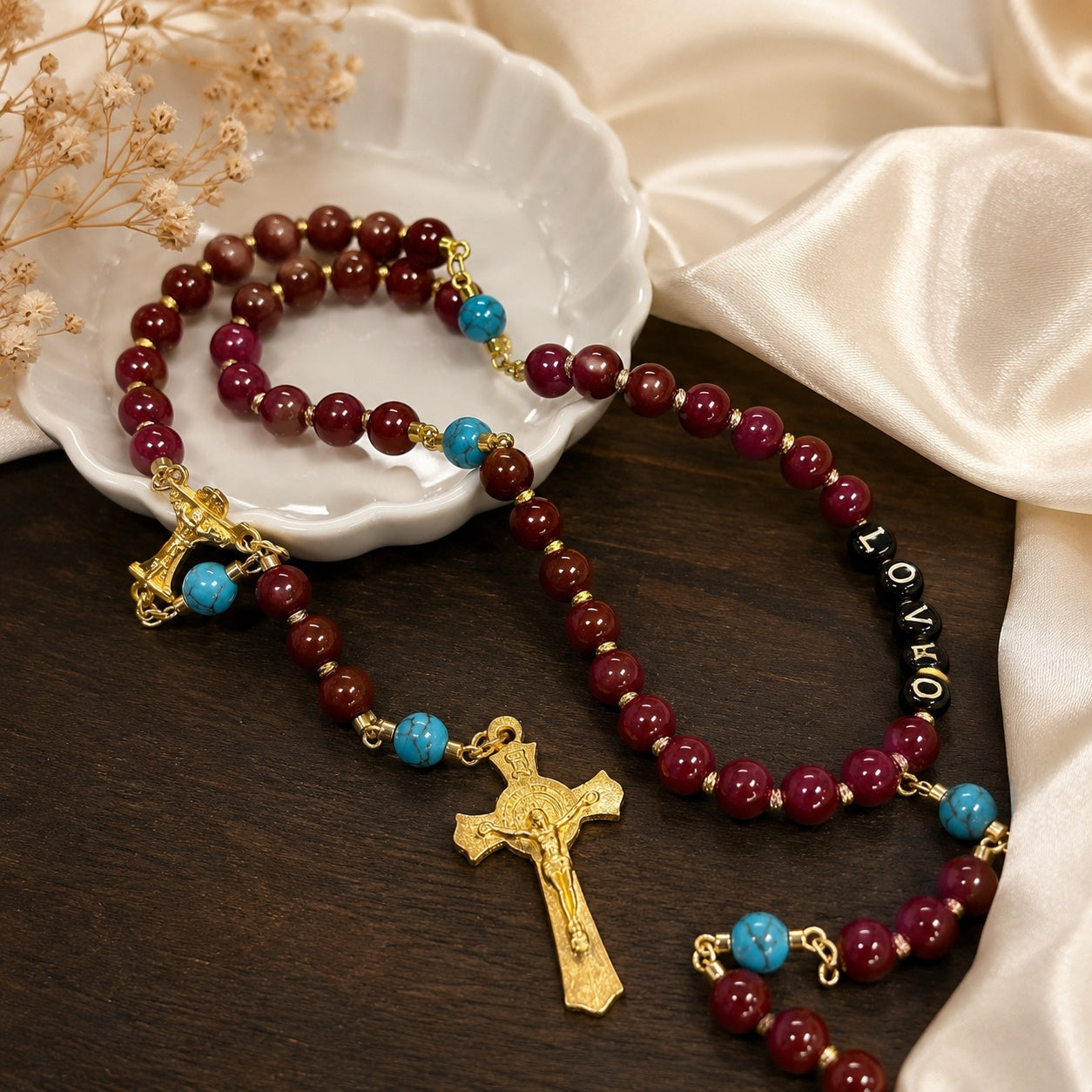 Red Catholic rosary with gold crucifix, turquoise accent beads, and LOVE letter beads displayed on a white ceramic dish