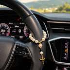 Pearl car prayer beads displayed in a white dish beside a candle and Christian decor, featuring a gold crucifix and initial bead